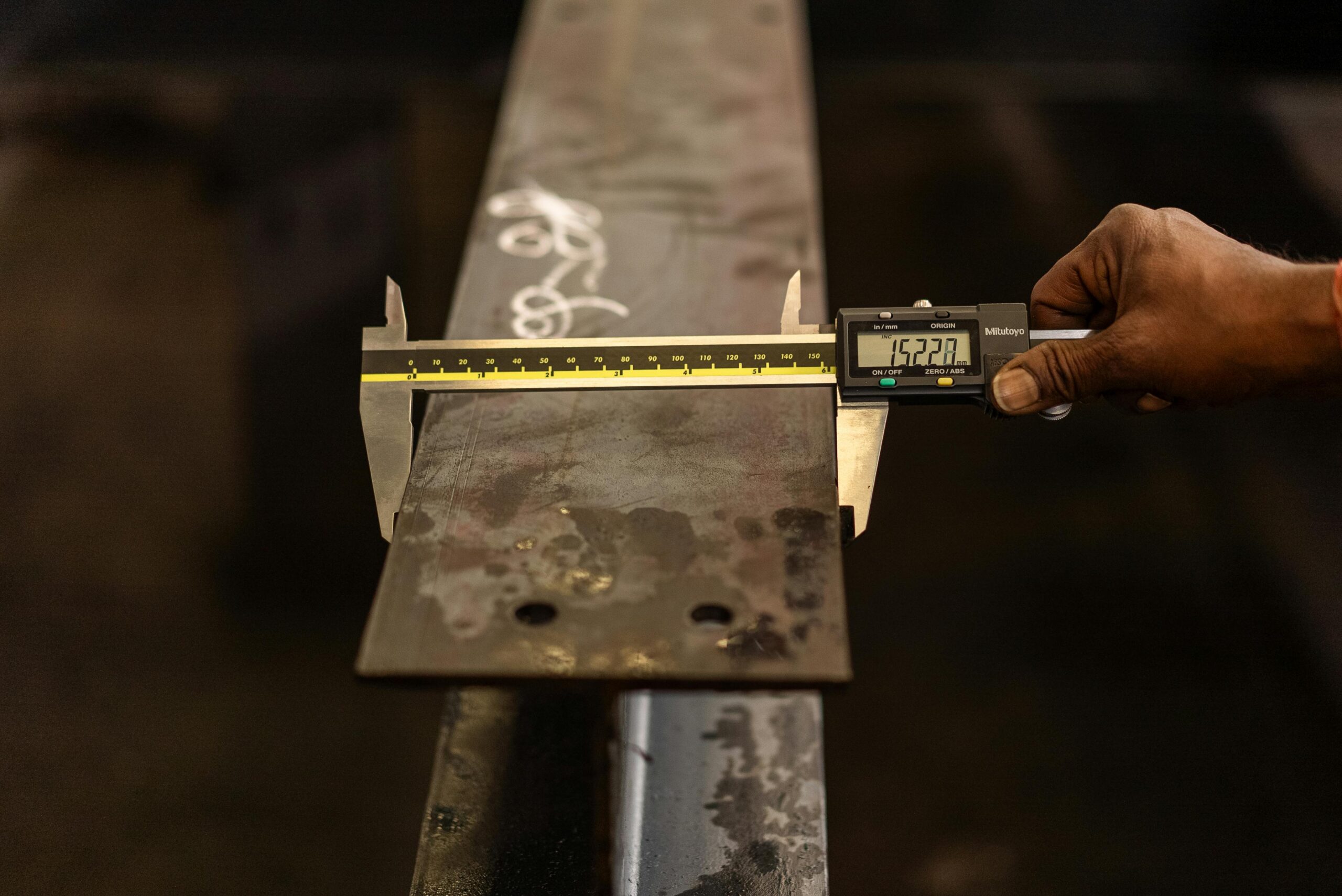 Close-up of a worker measuring a steel beam with a digital caliper in an industrial setting.
